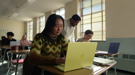 Student using a yellow Apple MacBook Neo laptop in a classroom while a teacher assists nearby.